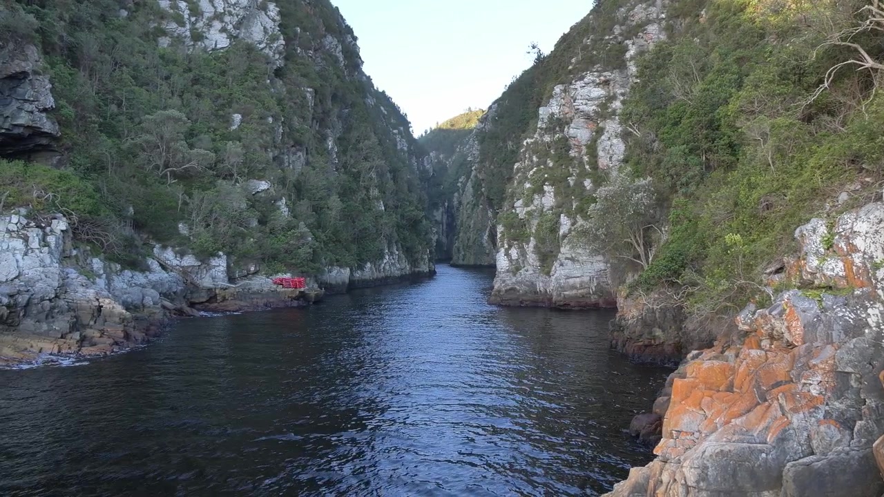 Ripresa slider da un ponte sospeso rivela un fiume tranquillo che si snoda tra imponenti scogliere verdeggianti, formazioni rocciose frastagliate, con kayak rossi impilati alla foce del fiume Storm, Tsitsikamma.