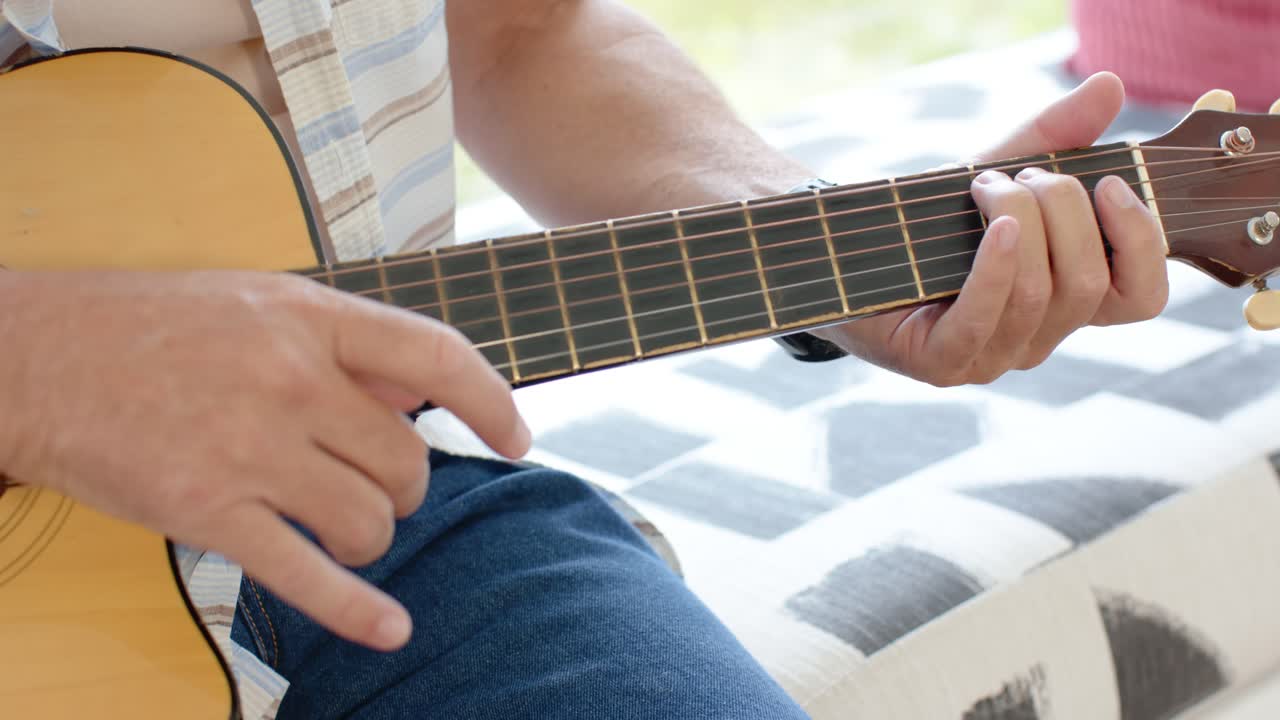 Hombre comenzando el rasgueo con la derecha, moviendo la mano izquierda y punteando una guitarra de corte junto a la ventana, interpretando una canción