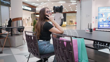 woman engaged in phone call, relaxing hand on table in modern urban setting with blurred background featuring shopping bags and contemporary structures