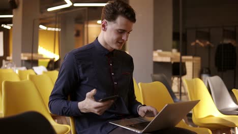 Young-Attractive-Male-In-A-Cute-Shirt-Smiling-While-Holding-Smartphone-And-Typing-Something-On-His-Laptop-At-The-Same-Time