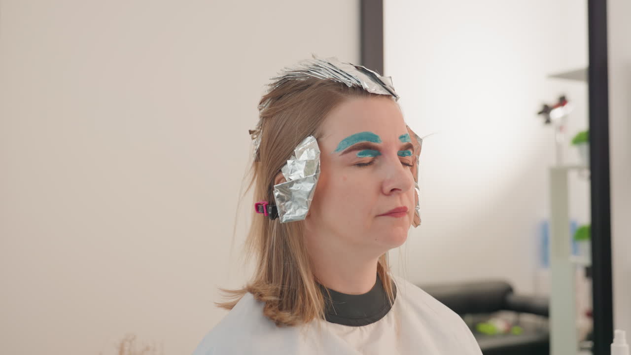 Kvinde i salon med foliestriber: Klienten sidder, mens frisøren påfører folie og farve, stylisten inddeler håret og fastgør folierne, makeupartisten lægger en markant blå øjenskygge, og en kappe er draperet over skuldrene.
