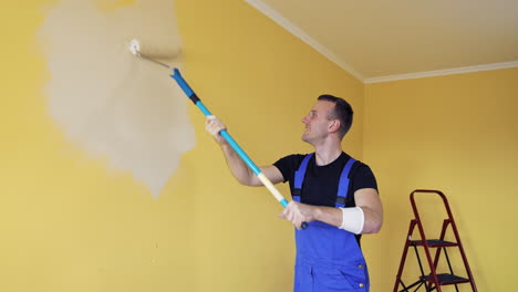 room improvement with a new color. professional worker in blue overalls painting wall with a paint roller on a long handle on. craftsman working.