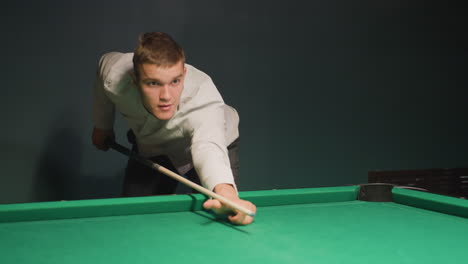 player in white shirt leans over green pool table, gripping cue stick with intense focus. eyes locked on cue ball as he prepares to take shot, demonstrating precision, skill, and concentration