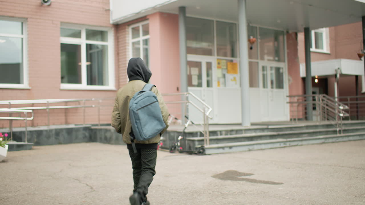 후드 재킷에 백팩을 메고 학교 건물 쪽으로 걸어가는 뒷모습의 남자아이, 계단 근처에 주차된 스쿠터, 흐린 아침 조용한 캠퍼스 입구, 도시 교육 풍경