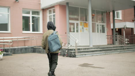 rear view boy in hoodie jacket with backpack walking toward school building, scooters parked near steps, quiet campus entrance on overcast morning, urban education scene