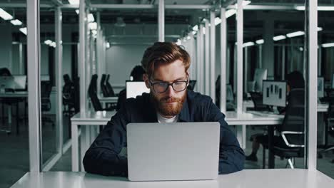 young businessman with eyeglasses and beard is working on laptop in modern office, focused on his tasks in a quiet and productive environment