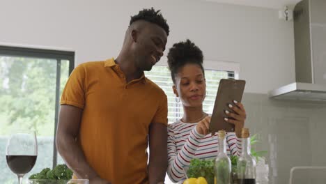 Video-of-happy-african-american-couple-cooking-together-in-kitchen,-using-tablet