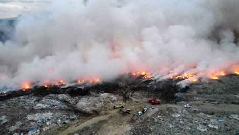 aerial, reverse, drone shot of a junkyard on fire, firefighter trucks in middle of garbage, waste and trash, toxic blue smoke rising, in santo domingo, dominican republic