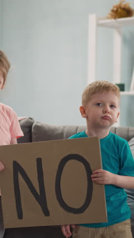 frowning little girl and boy holding poster with inscription NO, children are critical, in cozy room, protesting