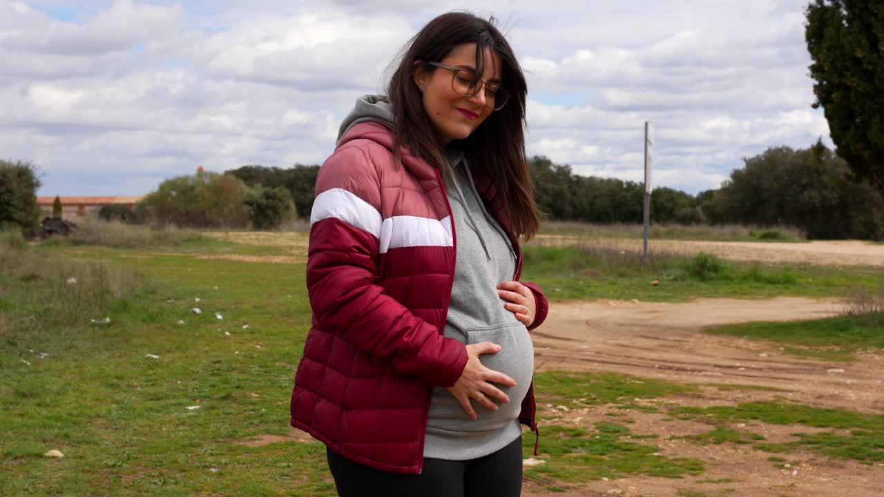 Happy pregnant woman smiles at camera while walking in open countryside on cloudy day