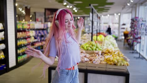 happy young girl with pink hair wearing wireless headphones listening to music and dancing in a grocery store