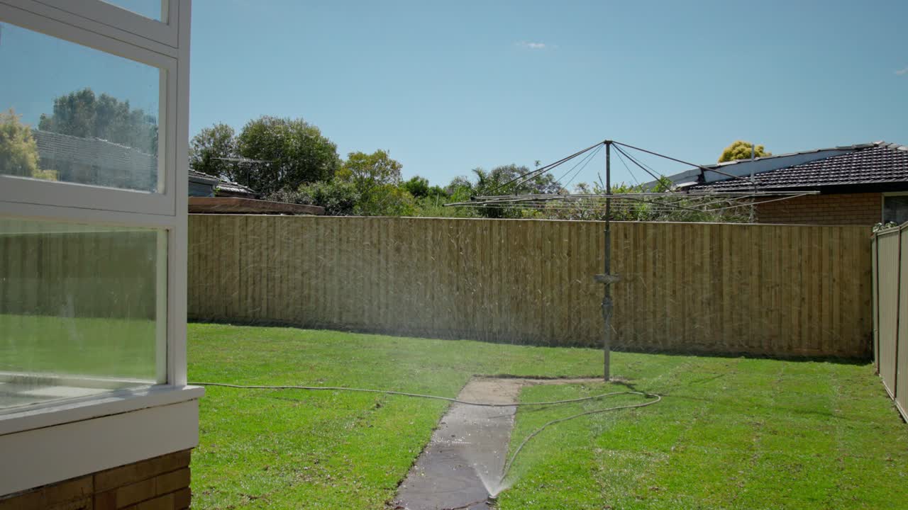 Static shot of a suburban residential backyard with a sprinkler actively watering a green lawn on a bright sunny day. A classic Hill Hoist rotary clothesline stands in the yard