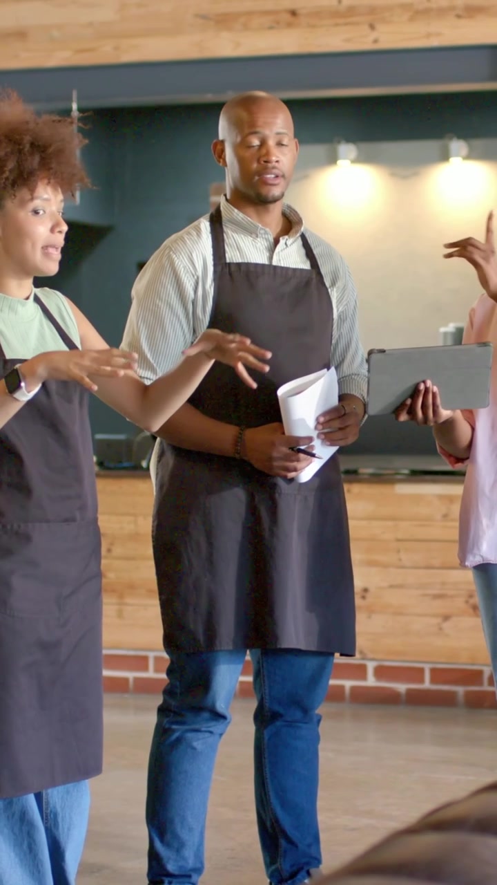 Vertikal video: Peger på tablet, får to medarbejdere til at planlægge layout på café med papirer, sorte forklæder