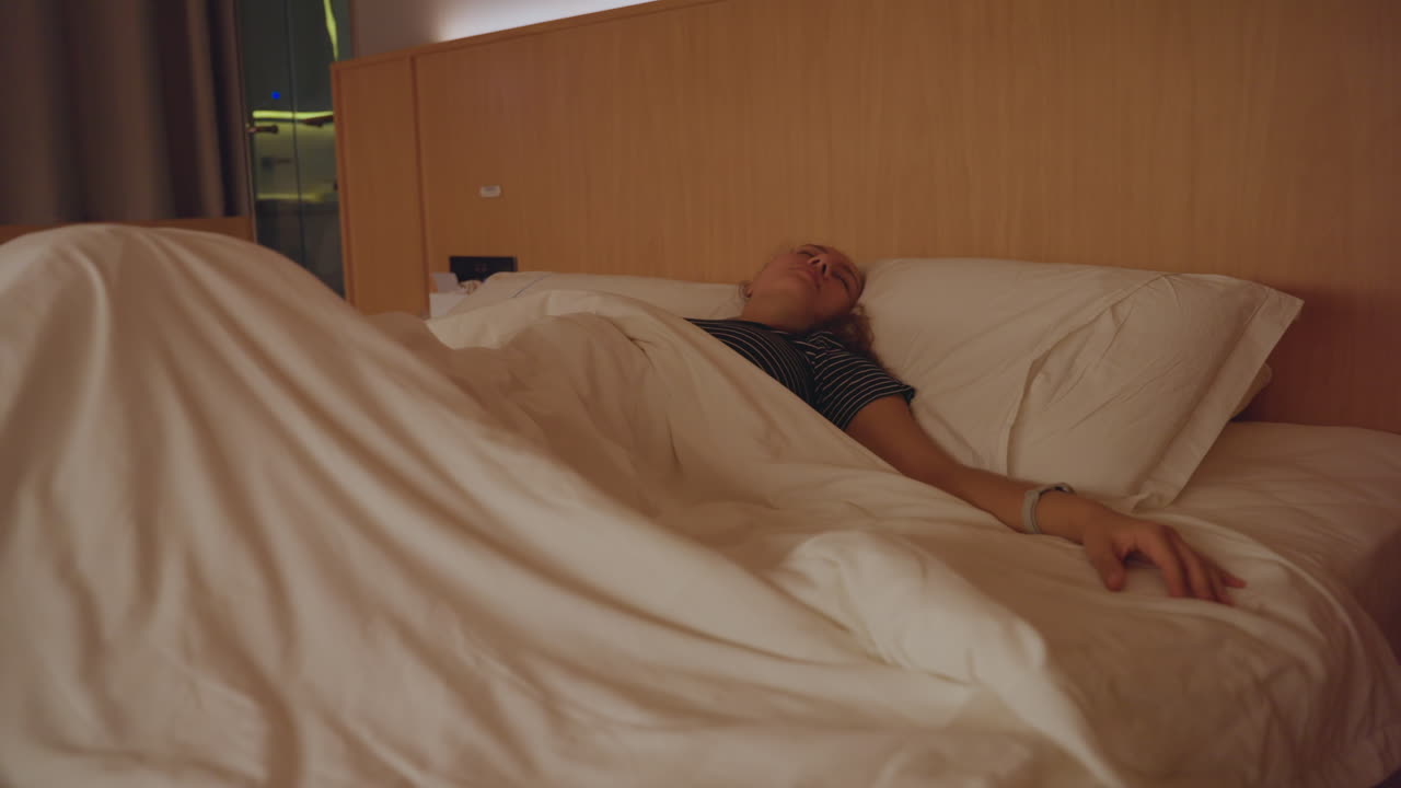 Hotelseng med en forretningsrejsende, der vågner og strækker sig efter et sent møde. Jakkesæt og slips er synligt på stolen, smartwatch på håndleddet, varm ravfarvet lampe, kompakt hotelværelsesinteriør og en stille morgenrutine.