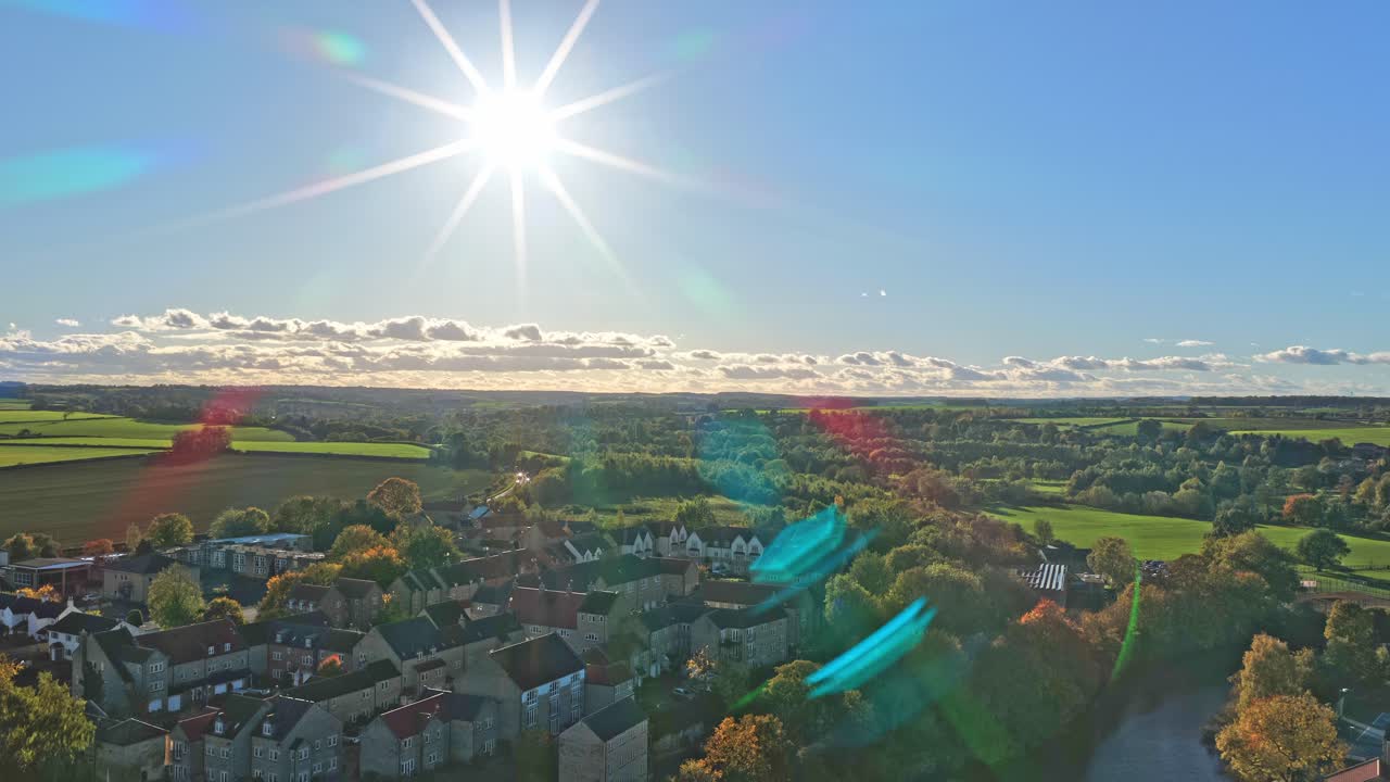 Elevated drone view unveils farmland, tree rows, open fields, residential rooftops, and sun rays dazzling above the outskirts of Wetherby, West Yorkshire, England