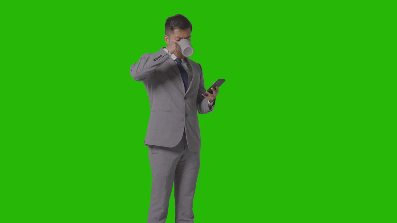Studio Shot Of Businessman In Suit Holding Hot Drink Looking At Mobile Phone Against Green Screen 