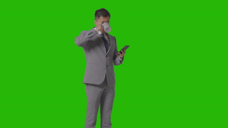 studio shot of businessman in suit holding hot drink looking at mobile phone against green screen