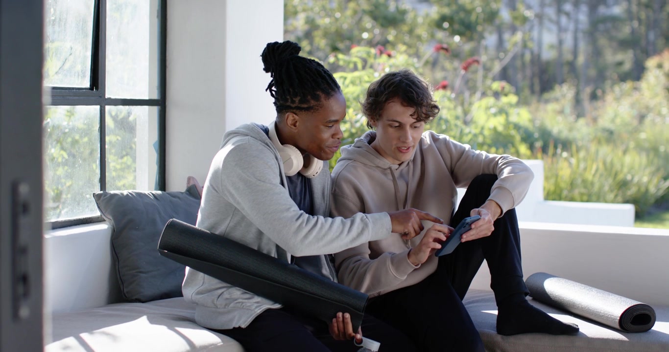 Two male friends sitting on sunny bench in hoodies, right man scrolling phone, planning workout