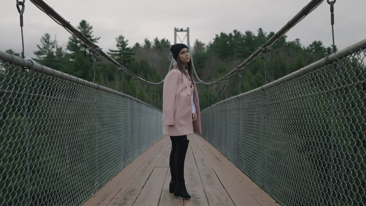 Skøn kvinde i pink blazer og sort bonetehat ved hængebro over Coaticook-floden, Eastern Townships, Quebec, Canada. Slowmotion-optagelse