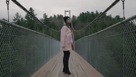 Encantadora-Mujer-Con-Chaqueta-Rosa-Y-Sombrero-De-Bonete-Negro-En-El-Puente-Colgante-Sobre-El-Río-Coaticook,-Municipios-Del-Este,-Quebec,-Canadá,-Tiro-En-Cámara-Lenta