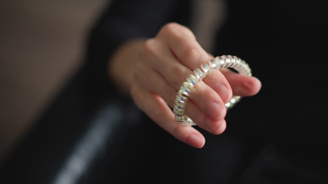 Primo piano di una mano che tiene e manipola un elastico per capelli a spirale iridescente su sfondo scuro. La mano si muove leggermente per mostrare l'accessorio flessibile, enfatizzandone la texture lucida e la forma. Morbido