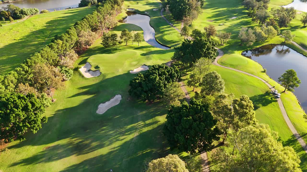 Drone footage captures a scenic golf course with lush greenery, sand bunkers, and water hazards under bright sunlight