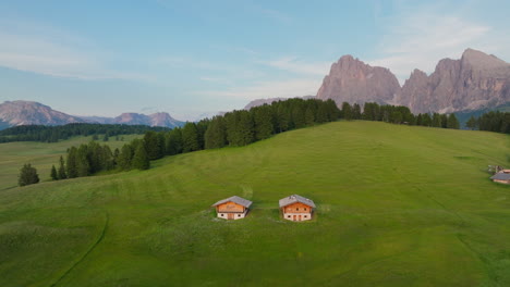 alpine wooden huts on alpe di siusi high altitude plateau with dolomites alps in background at sunset, panoramic view, drone shot