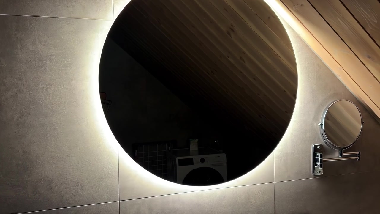 La telecamera si avvicina lentamente a uno specchio rotondo illuminato accanto a un piccolo specchio ingranditore in un accogliente bagno in mansarda