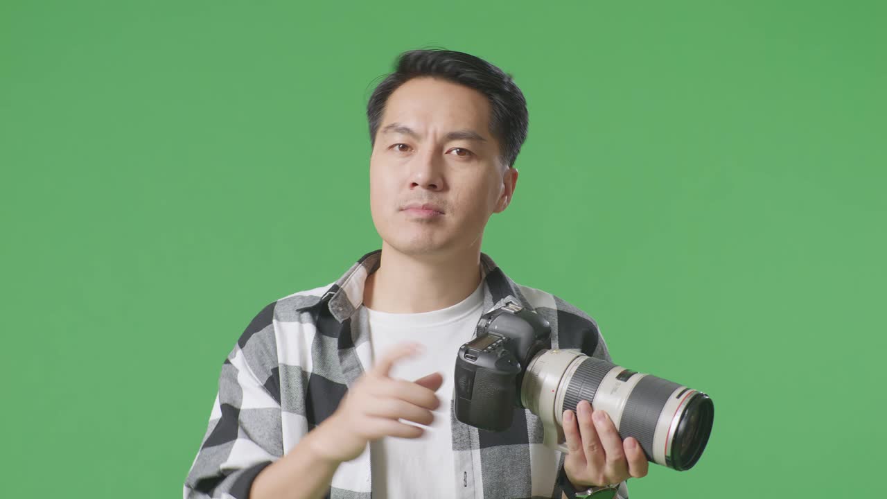 심각한 아시아 사진작가, 손에 카메라를 들고 스튜디오에서 녹색 화면 배경에 서있는 동안 지표손가락 표시 없이 불쾌감을 표합니다.