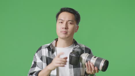 close up of serious asian photographer holding a camera in his hands and disapproving with no index finger sign while standing on green screen background in the studio