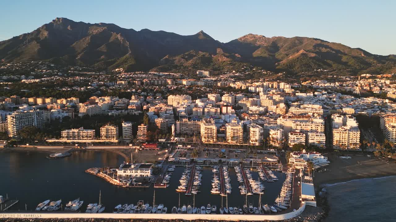 Vista aerea panoramica sulla splendida città e sul porto turistico di Marbella, Andalusia, Spagna, bagnati dalla luce dorata del tramonto e con le maestose montagne della Sierra Blanca come sfondo
