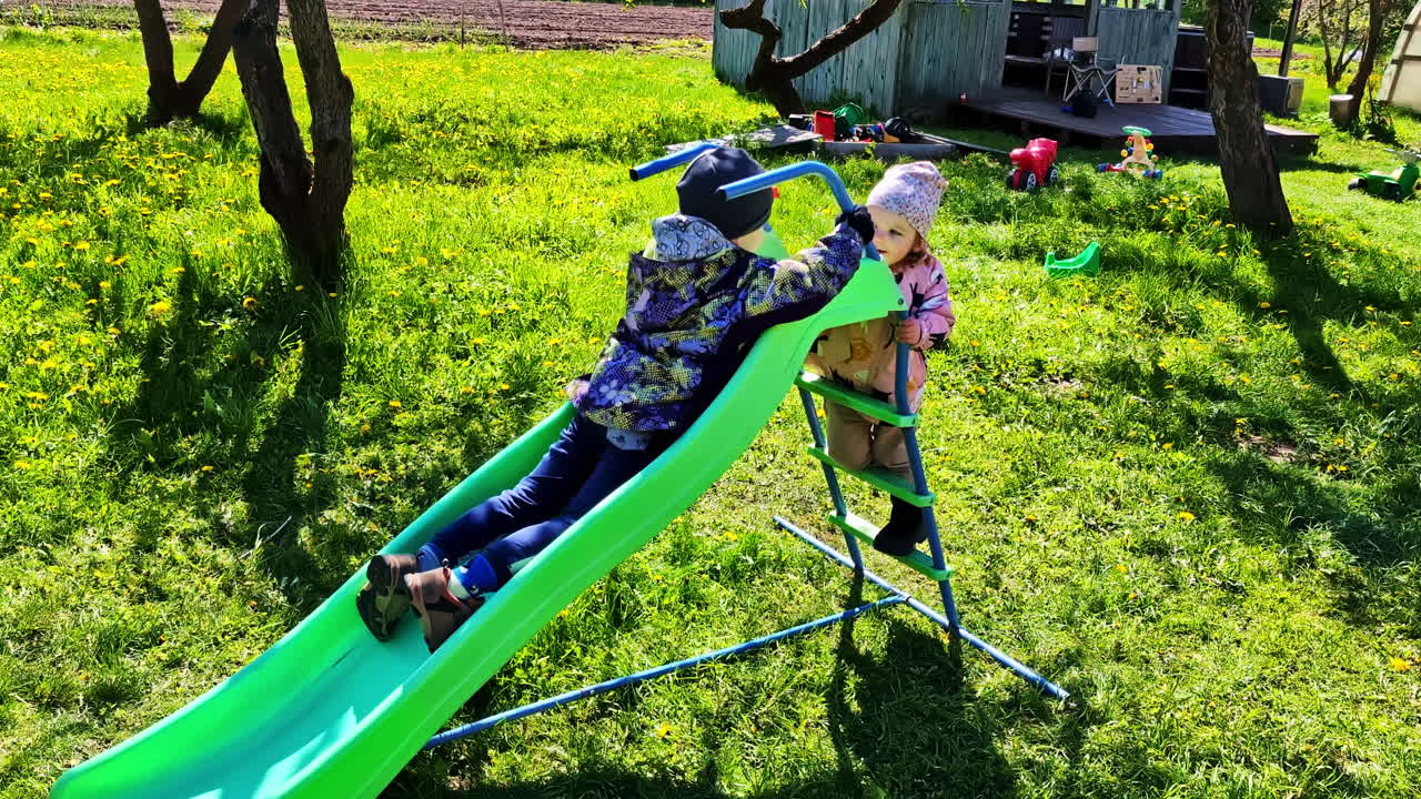 Young siblings climbing green slide ladder together in garden near wooden playhouse