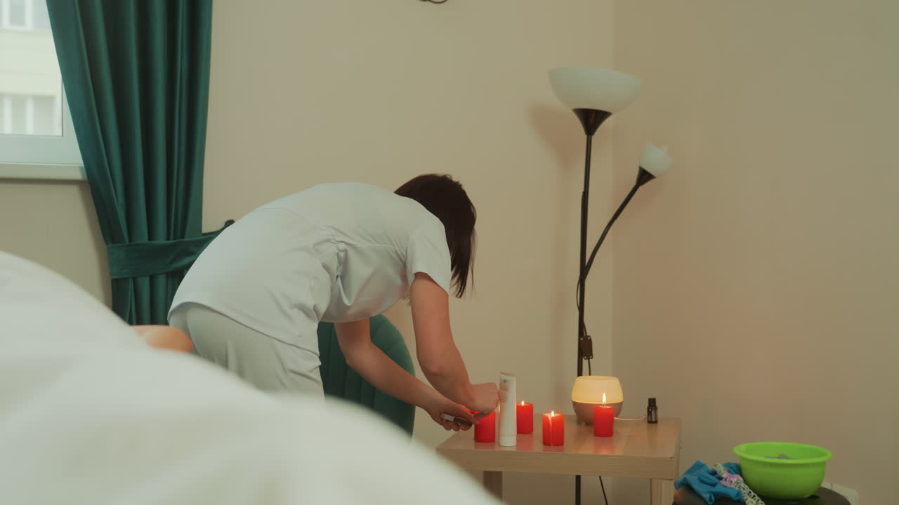 Terapis menyiapkan lilin untuk pijat mikrok arus di ruang perawatan yang tenang, menata minyak, lampu, handuk, mangkuk, dan seprai di samping tempat tidur pijat, praktisi memeriksa peralatan dan menciptakan suasana ambien yang menenangkan.
