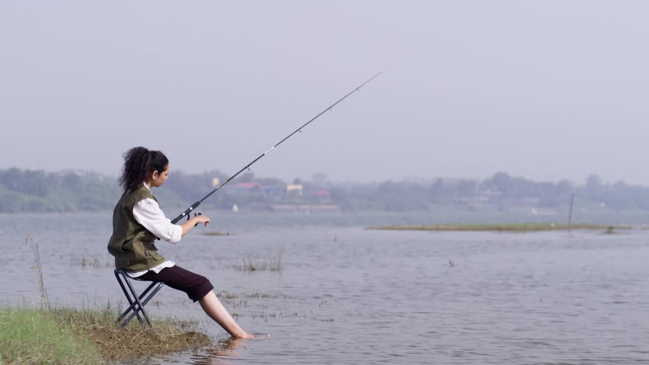 Seorang gadis Asia Selatan mengenakan rompi luar ruangan memancing dengan tenang sambil duduk di dekat air dangkal di danau alami yang tenang