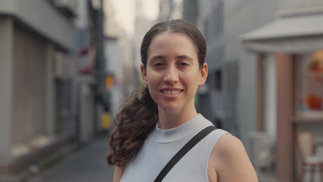 Ritratto di una giovane donna felice con i capelli ricci che sorride alla telecamera mentre si trova in uno stretto vicolo urbano a Tokyo, Giappone. Una bella turista che si gode la sua vacanza estiva all'estero.