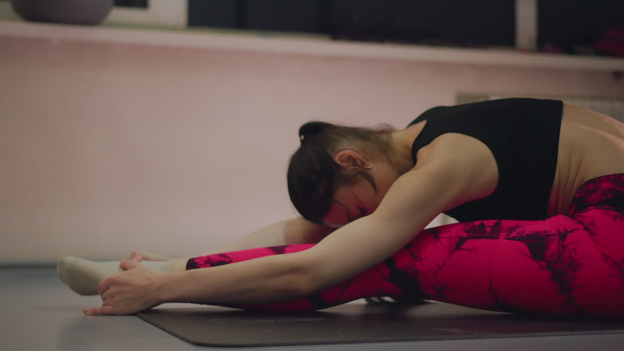 Donna bianca che esegue un piegamento in avanti profondo, rilascio degli ischiocrurali su tappetino in uno studio silenzioso, leggings e calzini rosa, respirazione lenta e postura meditativa, istruttrice che guida la pratica individuale.