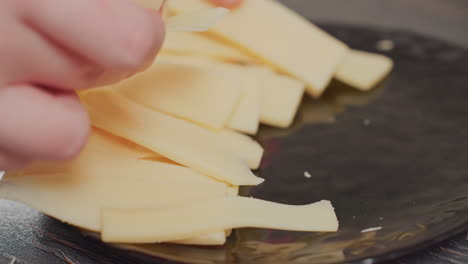 close up of person arranging sliced yellow cheese on black ceramic plate in kitchen with visible fingers gently placing pieces in natural soft light on rustic wooden surface with food crumbs
