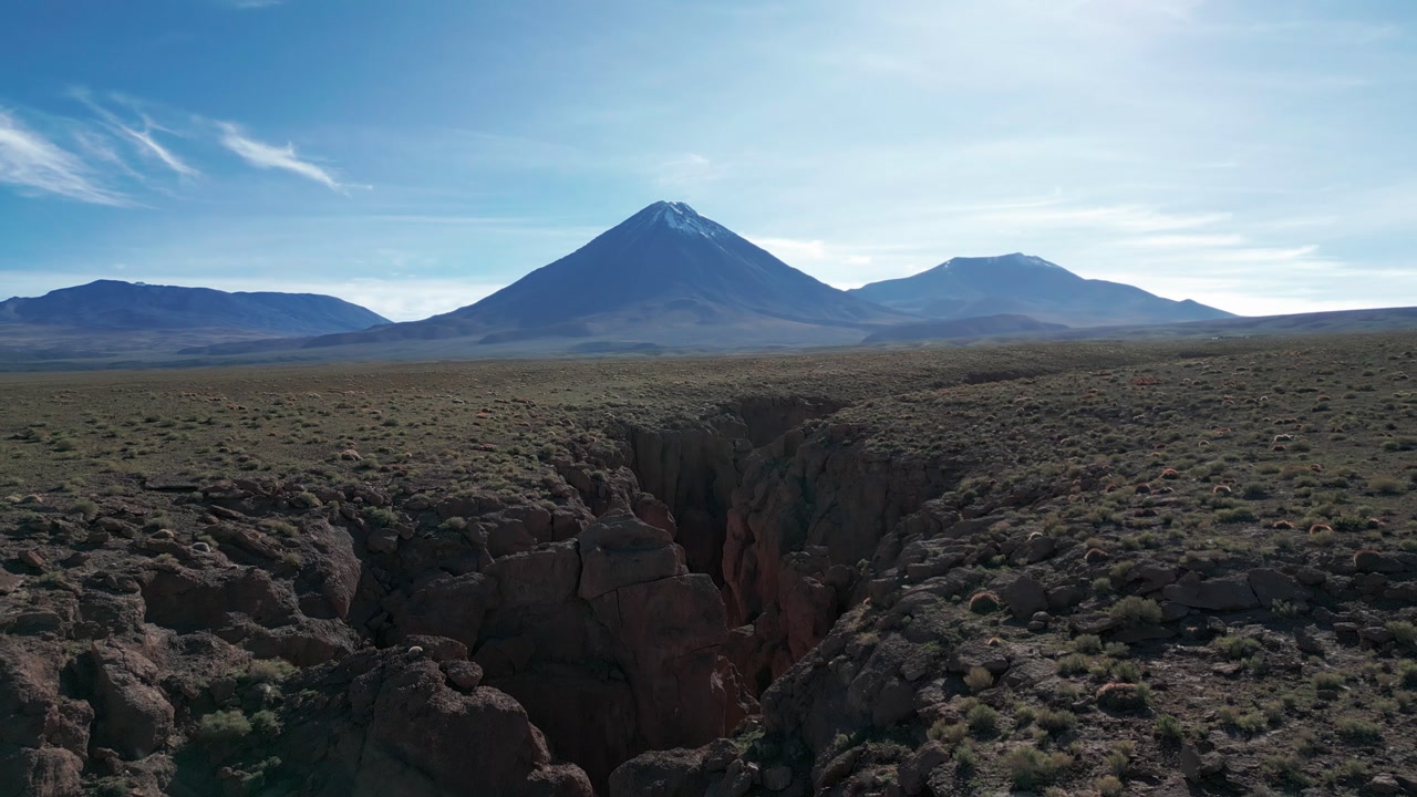 Backward drone flight over a dramatic rocky ravine revealing a vast arid altiplano plateau with Licancabur snow capped volcano dominating the horizon. San Pedro de Atacama, Chile