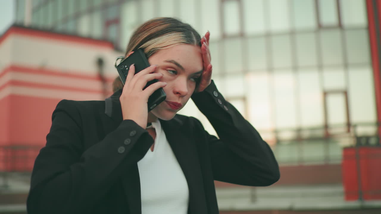 Lady in professional outfit appears tired while on phone call in modern urban setting, people blur in distant background, showing signs of exhaustion, stress, and emotional fatigue during conversation