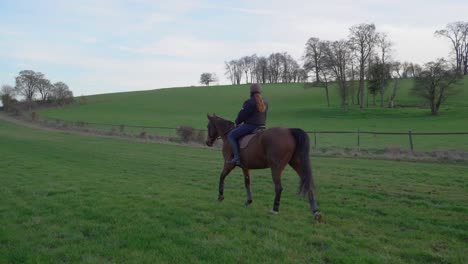 Young-girl-horseback-riding-a-brown-horse-on-a-green-lawn-in-the-countryside-on-a-sunny-day,-wide-shot