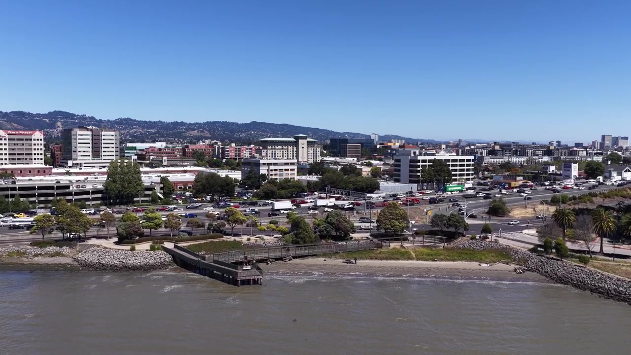 Drone che avanza sopra la costa della baia di Emeryville, CA, il molo, il parco sul lungomare e le strade trafficate, verso le colline di Oakland, California.