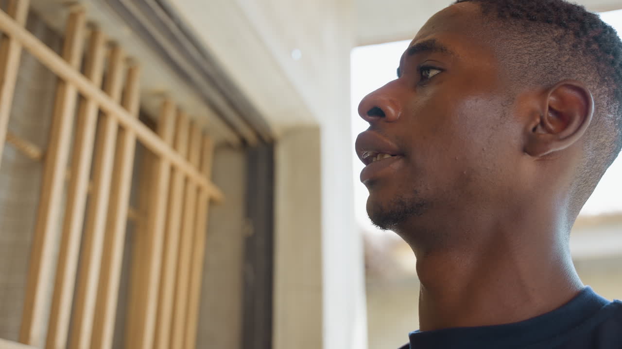 Black man repairing house window frame, sweaty handyman inspecting wooden slats, measuring and tightening screws inside sunlit residential interior, ladder and toolbox visible, focused expression