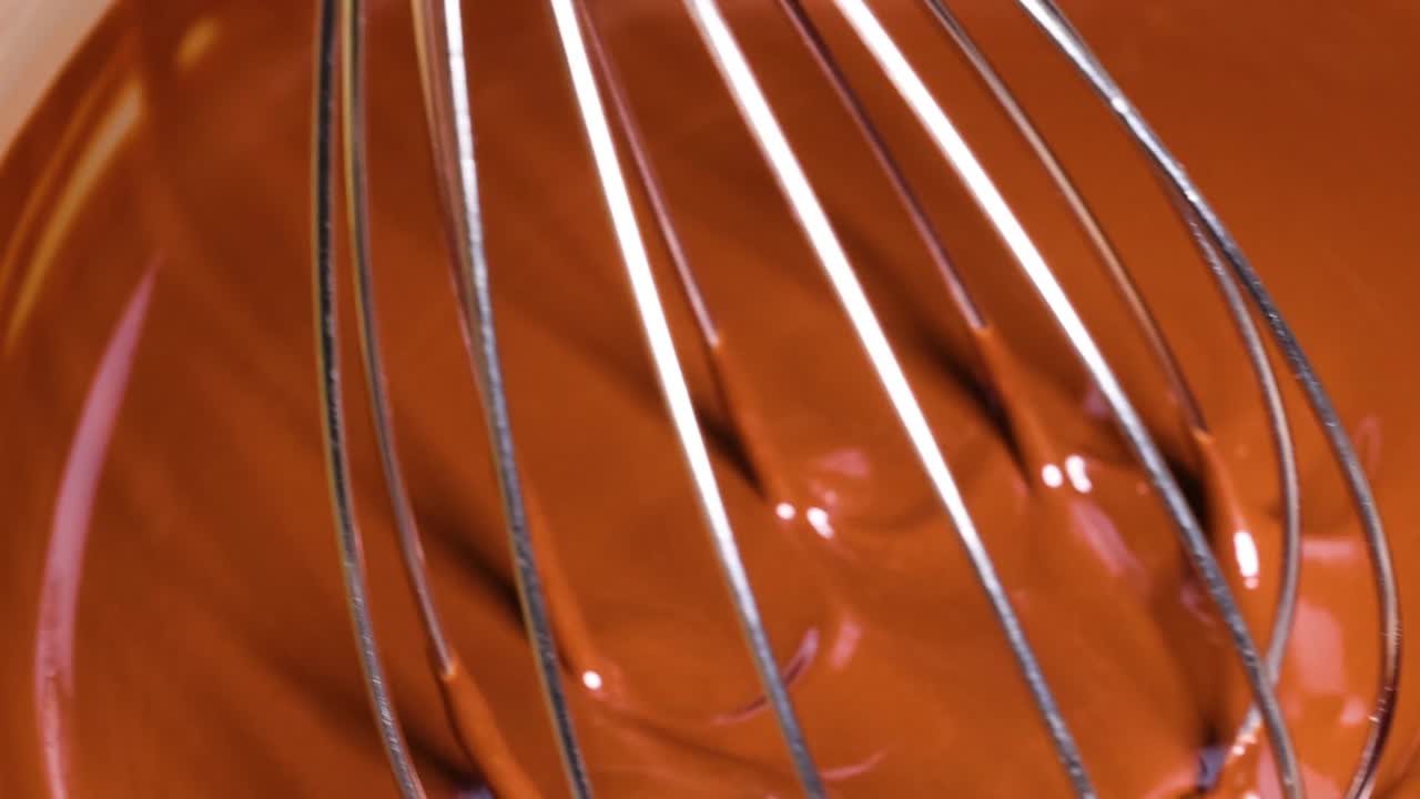 A detailed view of a metal whisk stirring smooth, melted chocolate, highlighting the glossy texture and swirling motion.
