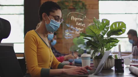 mujer caucásica con máscara facial usando una computadora portátil en una oficina moderna