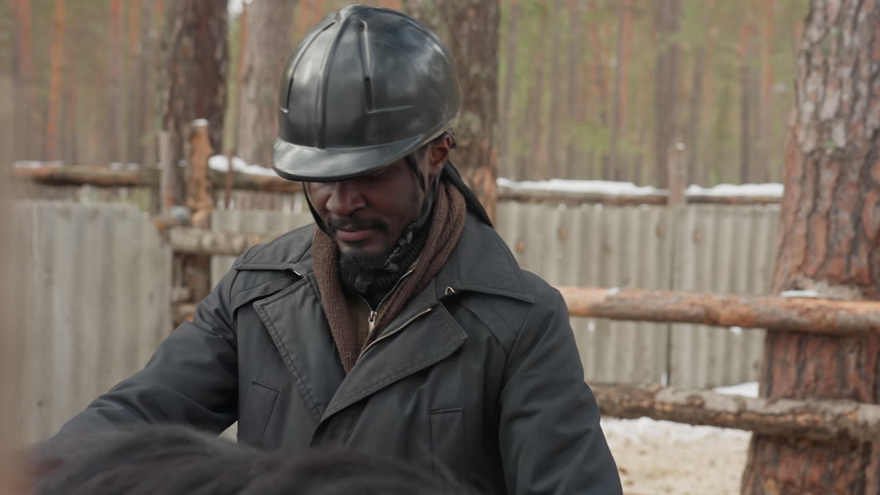 Un hombre revisa el equipo de un caballo; un hombre inspecciona el equipo en un establo; un individuo examina los arreos en un entorno de establo nevado; un hombre con casco revisa el equipo de un caballo dentro de un establo en invierno.