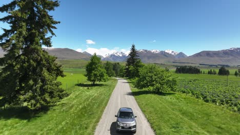 Car-driving-on-gravel-road-in-rural-New-Zealand-with-distant-mountains,-aerial