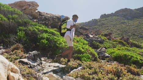 African-american-man-wearing-face-mask-exercising-outdoors-hiking-in-countryside-on-a-mountain