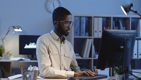 Office-Worker-Sitting-At-Desk-With-Computer,-Trying-To-Rest-While-Massaging-His-Neck-And-Taking-Off-His-Glasses-In-The-Office-At-Night