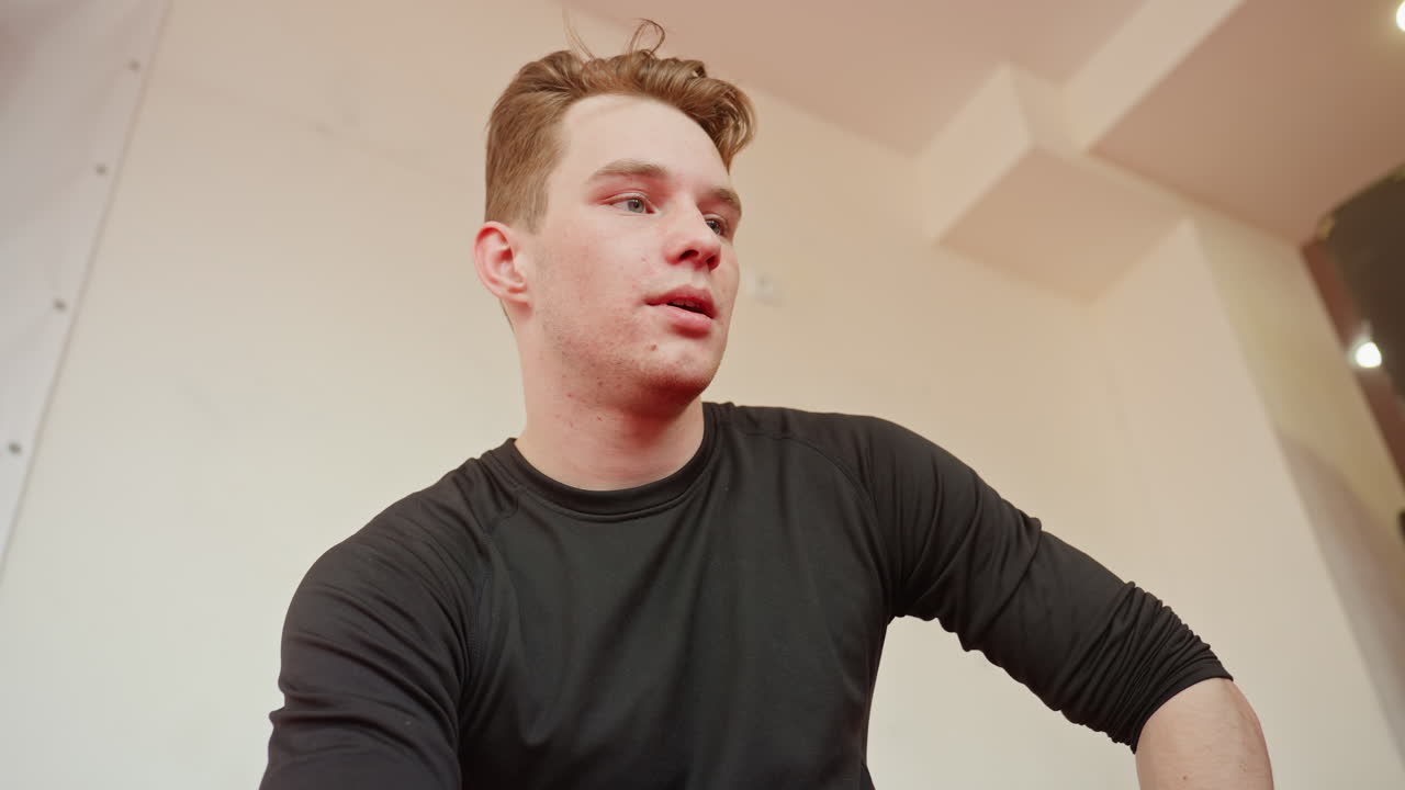 Male fighter resting after sparring in gym, wearing black sportswear, breathing heavily with tired yet focused expression, face showing fatigue, resilience under training lights after intense workout