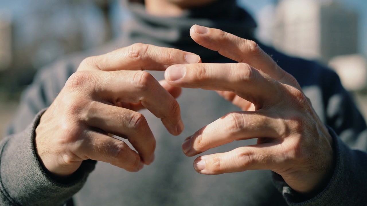 Rubbing man bringing fingers together after chill for warmth in sunlit city, grey hoodie dark scarf